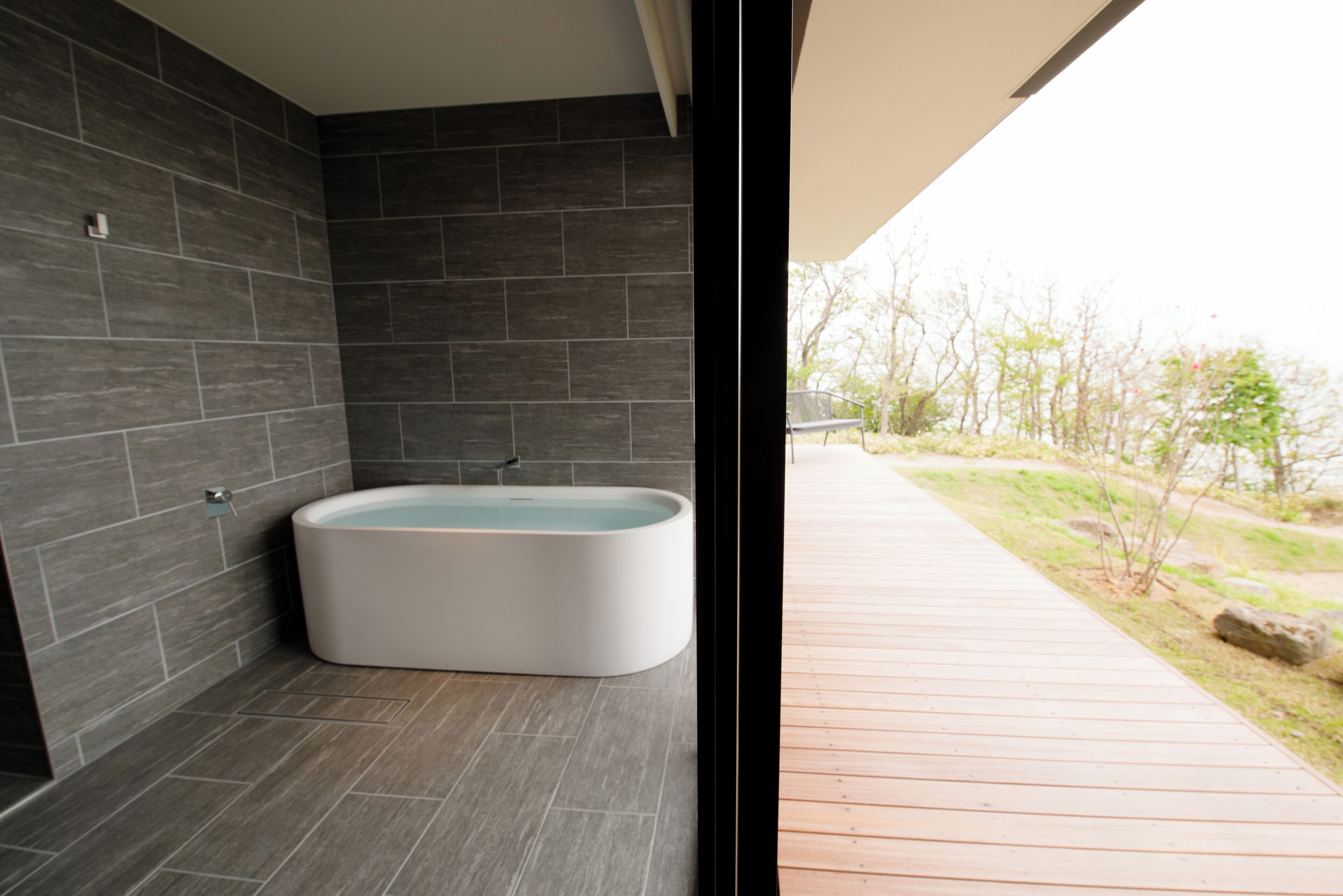 Une salle de bain moderne avec une baignoire îlot, des murs recouverts de carreaux foncés et une terrasse en bois donnant sur l’extérieur.