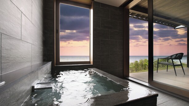 Un baño moderno con una bañera grande, vistas al mar a través de una puerta de cristal y una terraza de madera con una silla.