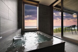 A modern bathroom with a large bathtub, a view of the sea through a glass door, and a wooden deck with a chair.