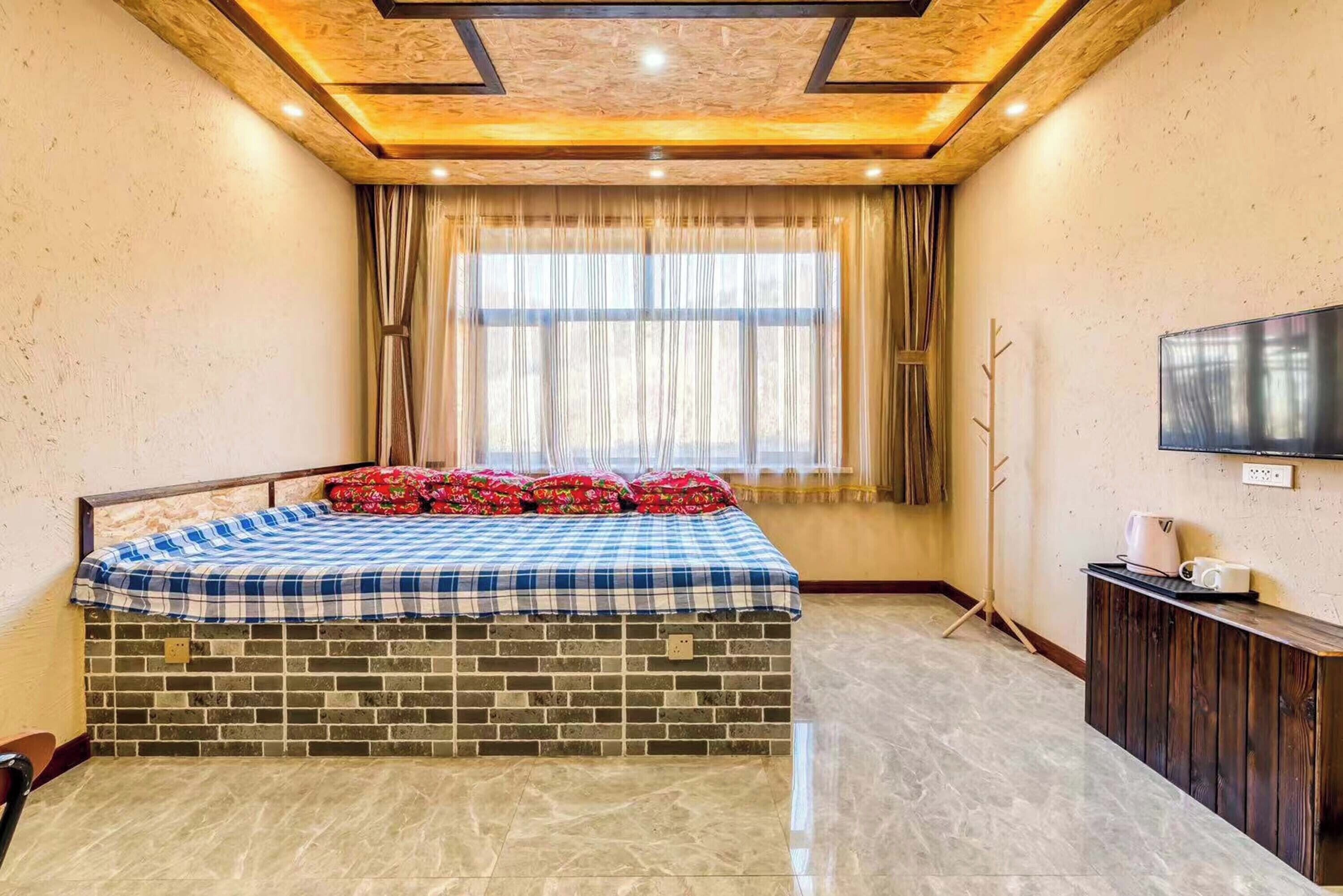 A bedroom with a brick-patterned headboard, a checkered bedspread, a flat-screen TV on the wall, and a window with curtains.