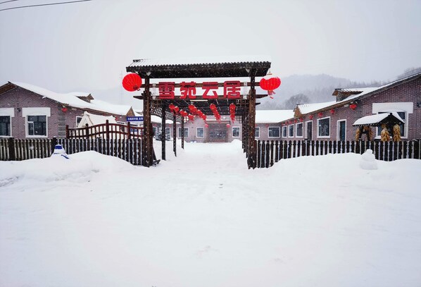 A traditional Chinese gate with red lanterns, leading to a row of brick buildings with snow on the ground.