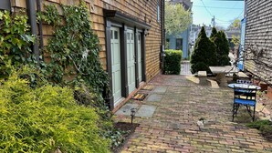 A narrow brick pathway leading to a wooden house with a green door, surrounded by greenery and a small table with chairs. - 86 Dr Marcus House - Popular Broadway Area (Newport)