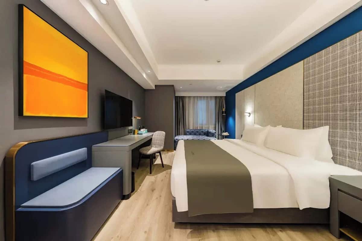 A modern hotel room with a bed, desk, chair, and a large abstract painting on the wall.