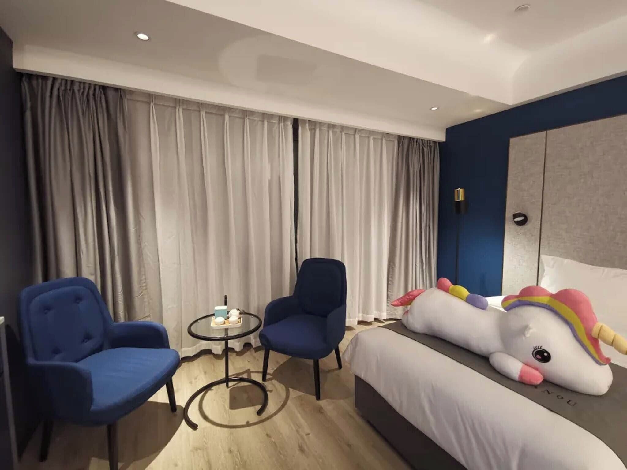 Une chambre d’hôtel moderne avec un grand lit, deux fauteuils bleus, une petite table ronde et une grande décoration représentant une licorne.