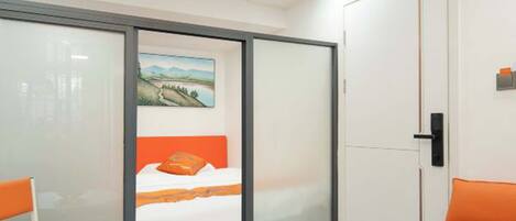 A modern hotel room with a bed, a dining table with fruit, and a bright orange chair.