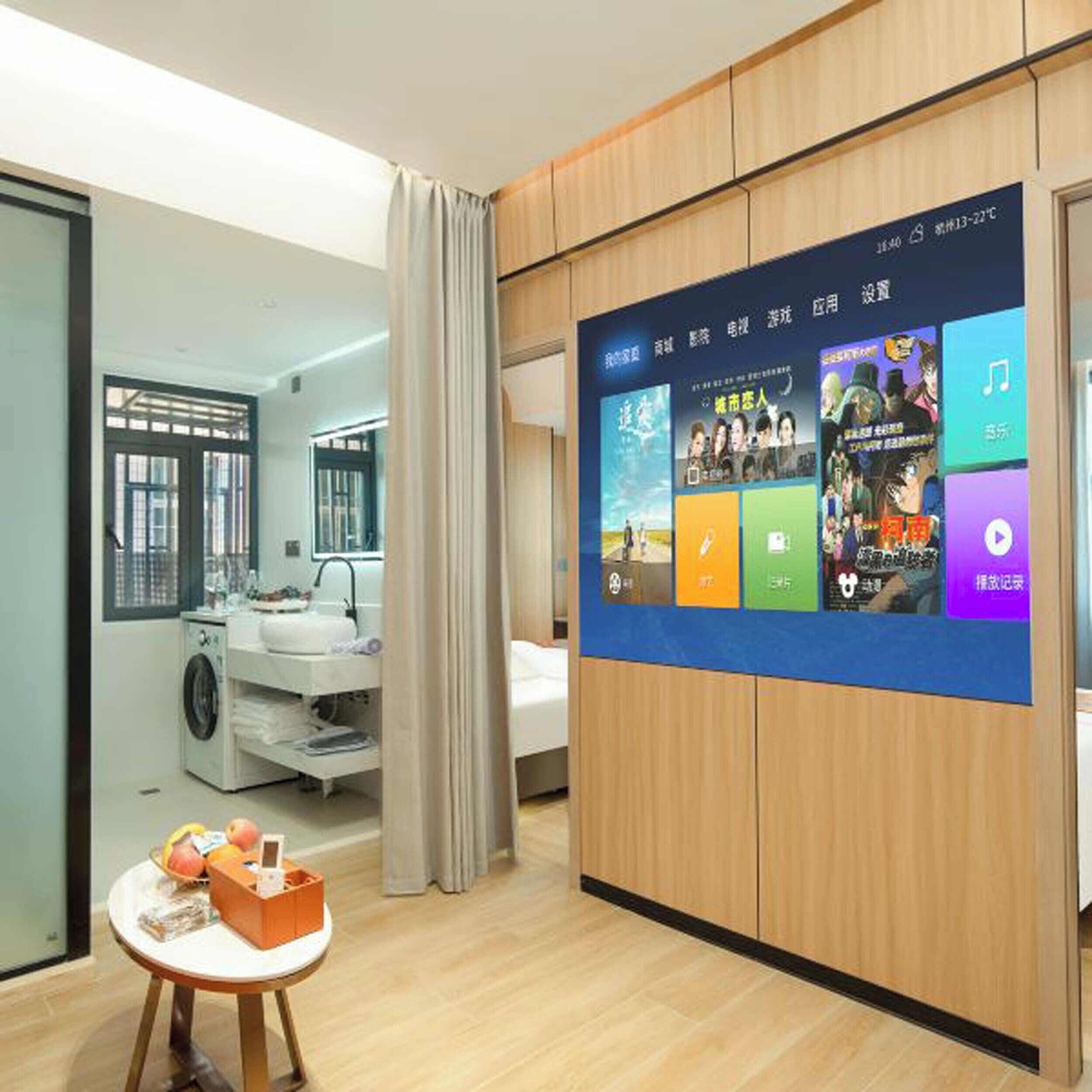 Hotellihuone, jossa on suuri digitaalinen näyttö seinällä, pyykinpesukone, pesuallas ja pieni pöytä, jonka päällä on hedelmiä ja pientä purtavaa.