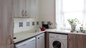 A compact kitchen with wooden cabinets, a washing machine, and a dishwasher. - Cosy 1-Bedroom Flat, Walk to Beaches (South Ayrshire)
