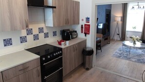 A modern kitchen with wooden cabinets, a black stove, and a tiled backsplash. There is a small trash can and a coffee maker on the counter. - Cosy 1-Bedroom Flat, Walk to Beaches (South Ayrshire)