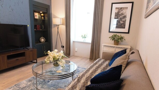 A living room with a sofa, a coffee table with flowers, a TV, and a bookshelf. - Cosy 1-Bedroom Flat, Walk to Beaches (South Ayrshire)