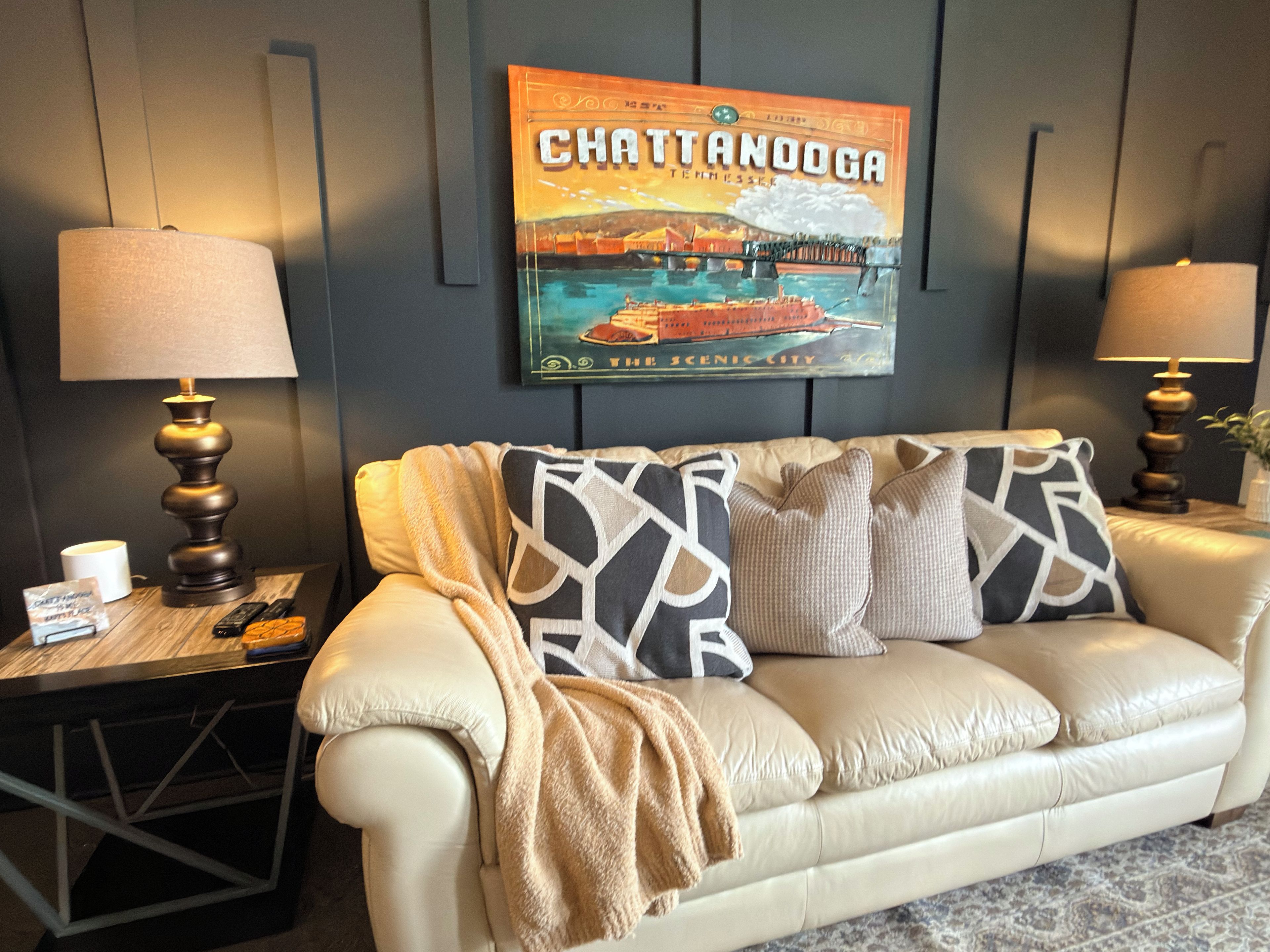 A room with a white leather sofa, two lamps, a framed picture of Chattanooga, and a small table with a remote control.