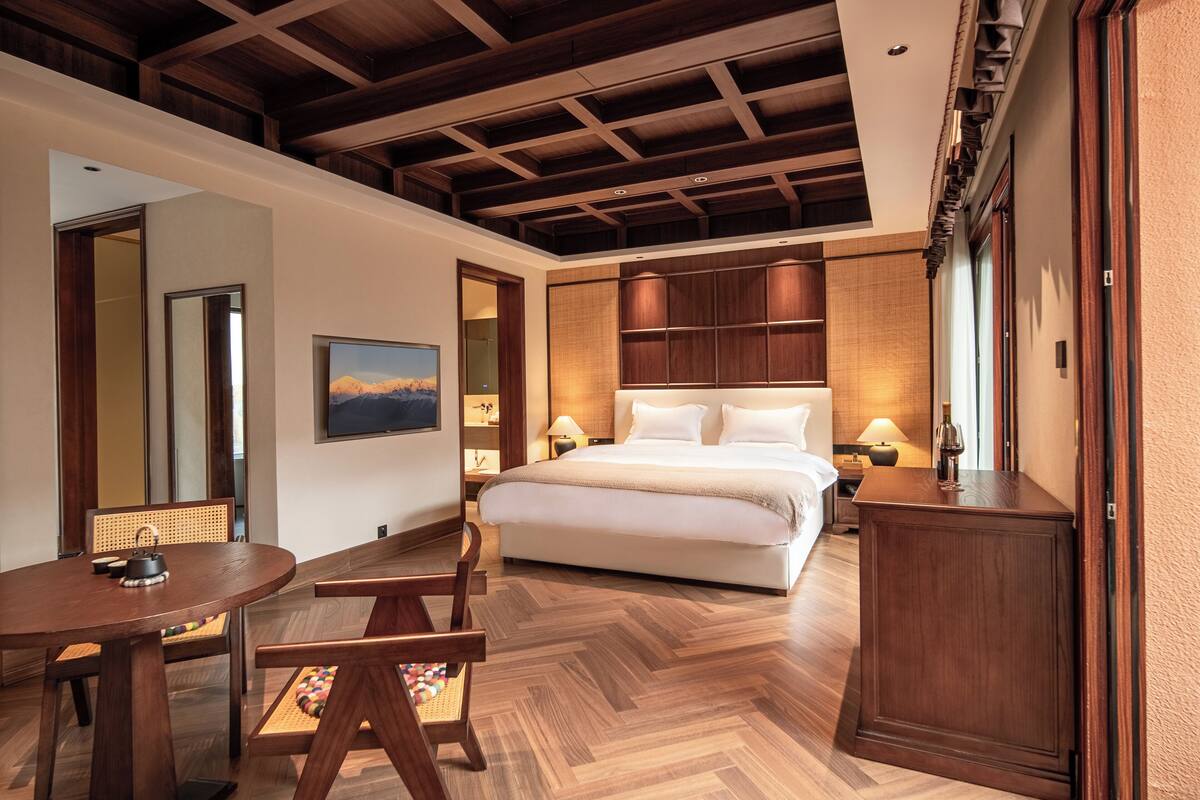 A hotel room with a large bed, wooden flooring, a dining table with chairs, a small kitchen area, and a large mirror.