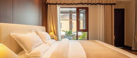 A neatly made bed with white linens and a beige throw blanket, a bedside lamp, and a view of the outdoors through a window with curtains.