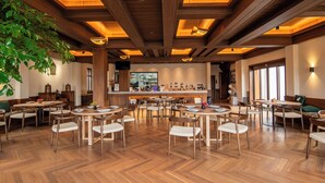 A spacious dining area with multiple round tables and chairs, wooden flooring, and a high ceiling with exposed beams and pendant lighting.