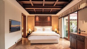 A bedroom with a large bed, wooden flooring, a ceiling with exposed beams, a nightstand with lamps, a painting on the wall, and a view through the window.