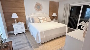 A bedroom with a bed, nightstands, a wooden side table, a ceiling fan, a large mirror, and a view of the outside through a sliding door.