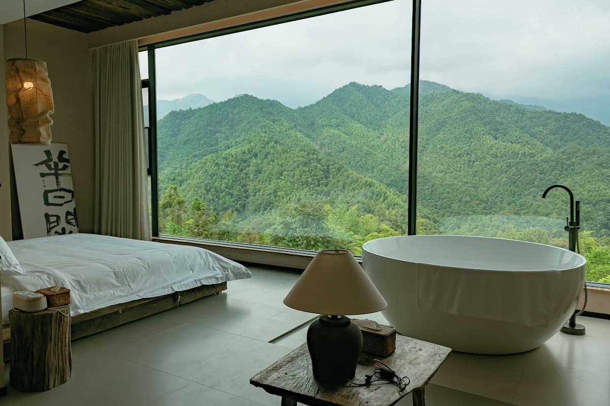 A bedroom with a large window, a bed, a bedside table with a lamp, and a large bathtub.
