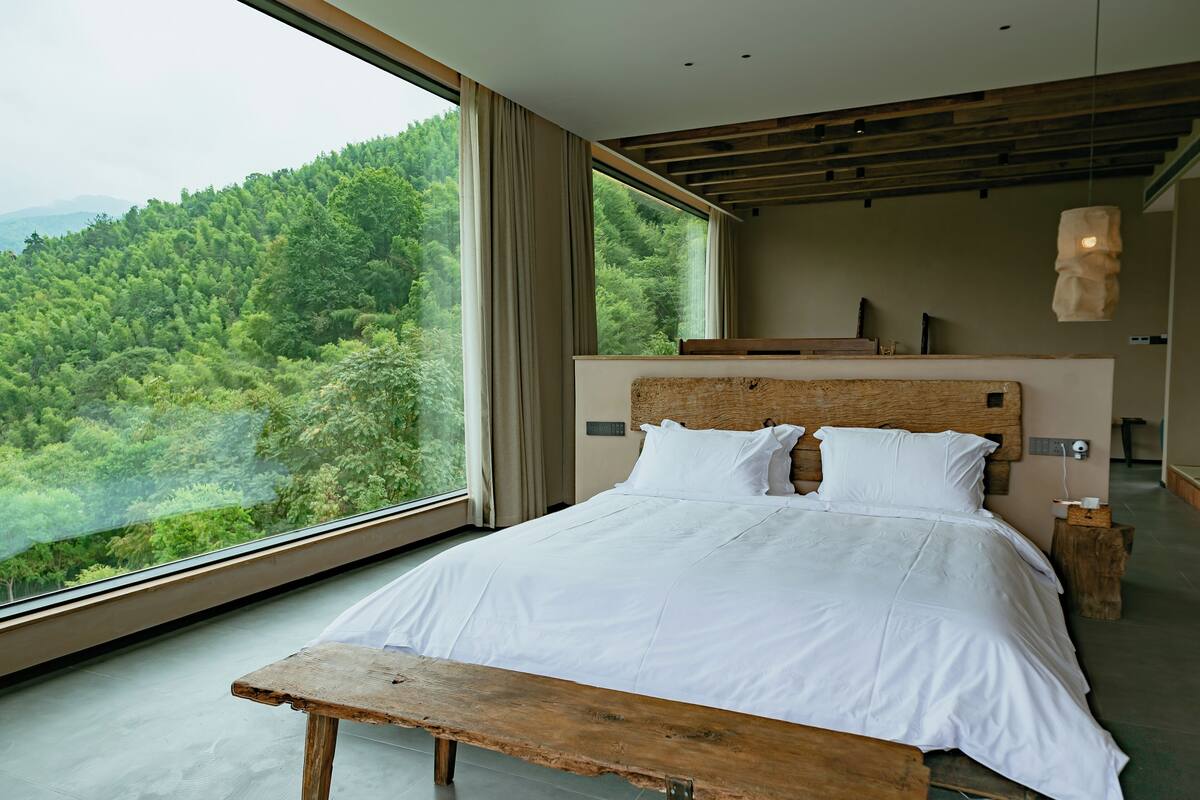 A bedroom with a large window, a wooden headboard, and a wooden bench.