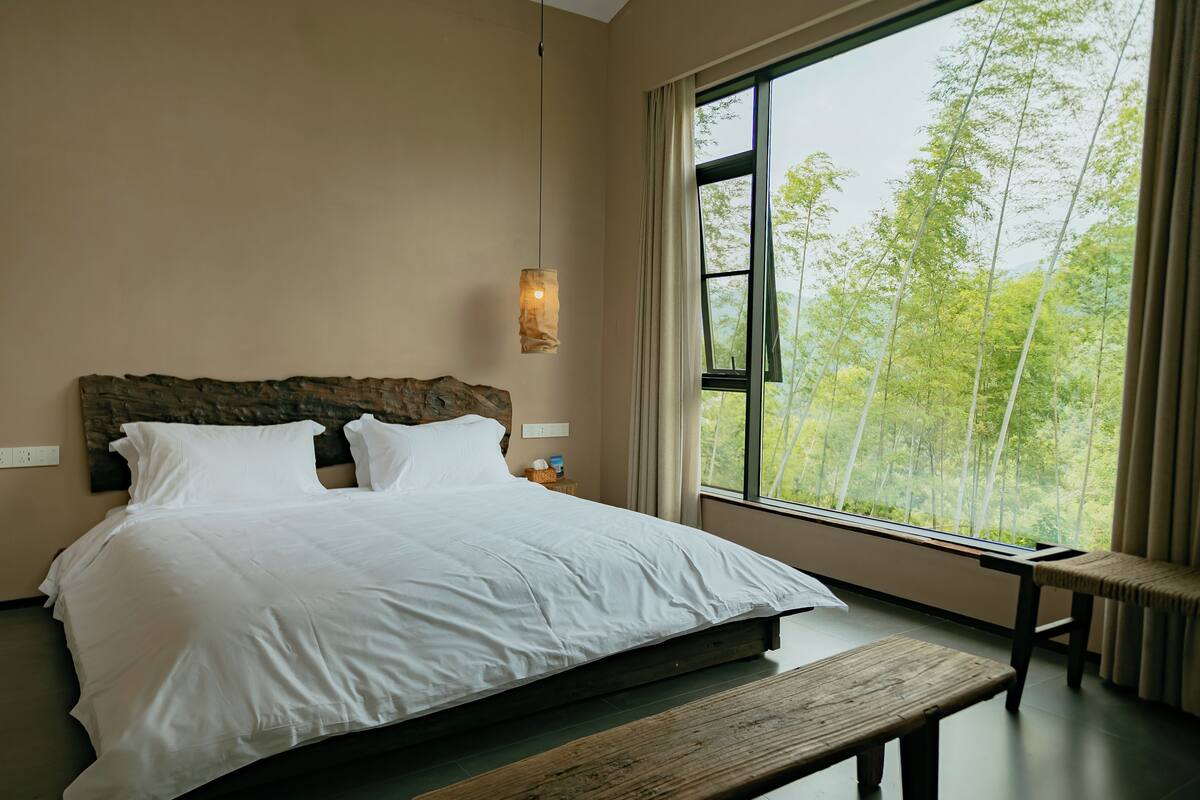 A bedroom with a large bed, a wooden headboard, a bedside table, a bench, and a window with a view of trees.