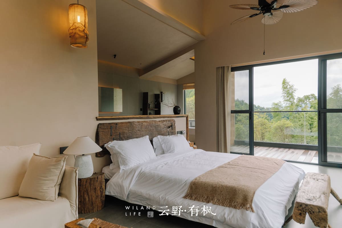 A bedroom with a large bed, a nightstand, a lamp, and a view of the outdoors through a large window.