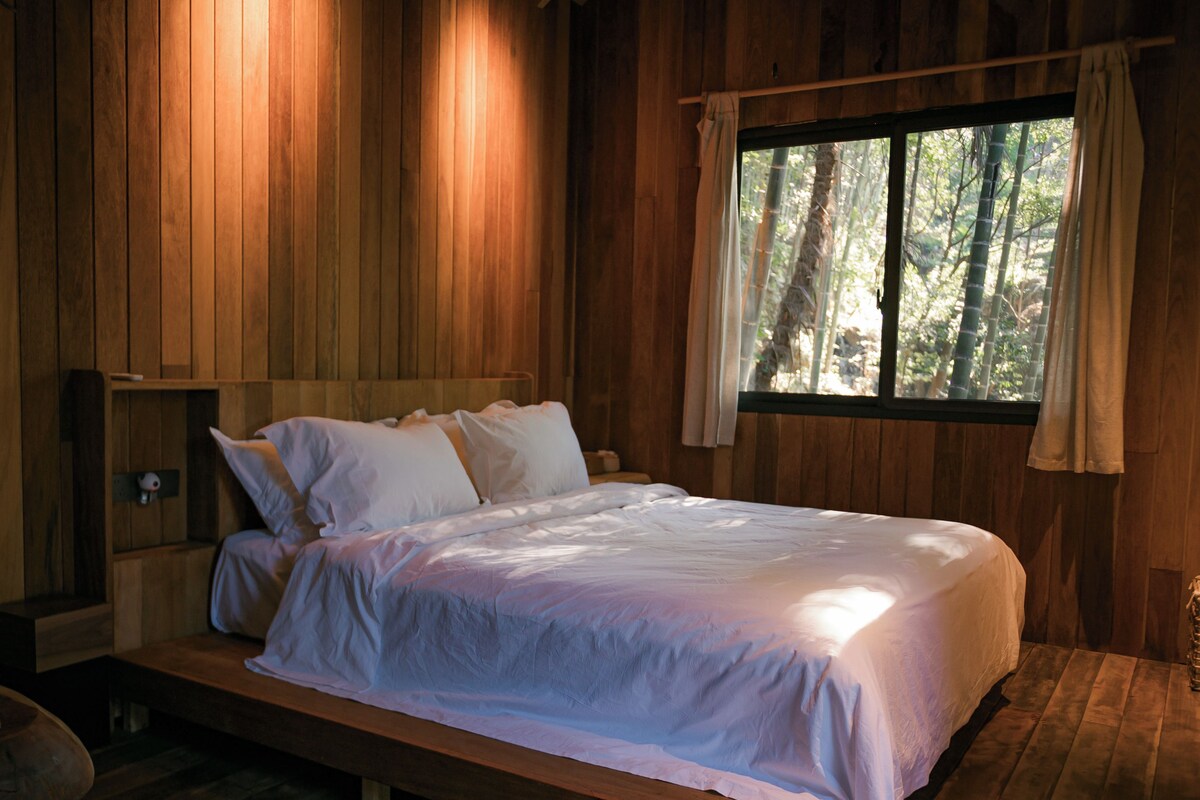 A wooden cabin bedroom with a bed, a window with curtains, and a small wooden bench.