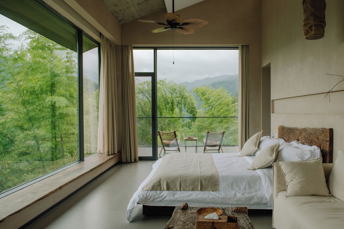 A bedroom with a large bed, a sofa, and a view of greenery through the window.