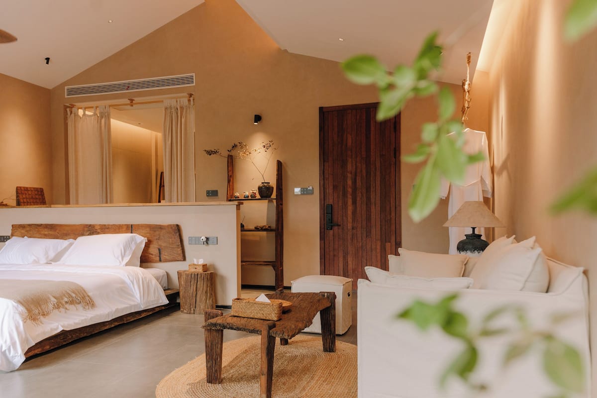 A bedroom with a large bed, a wooden bench, a small table, a sofa, a mirror, and a wooden wardrobe.