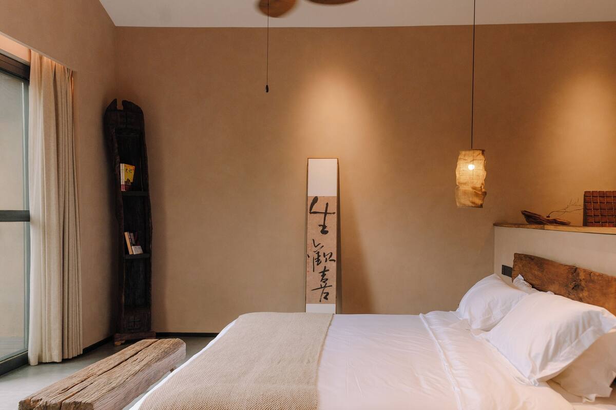 A bedroom with a large bed, a wooden headboard, a bedside table, a hanging light fixture, and a wall-mounted artwork.