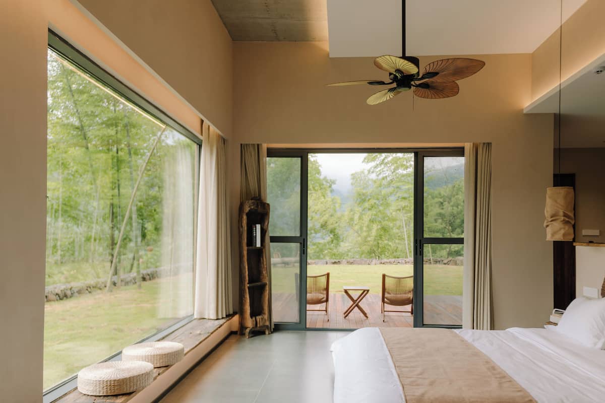 A modern bedroom with a large bed, a ceiling fan, and a view of a garden through a sliding door.