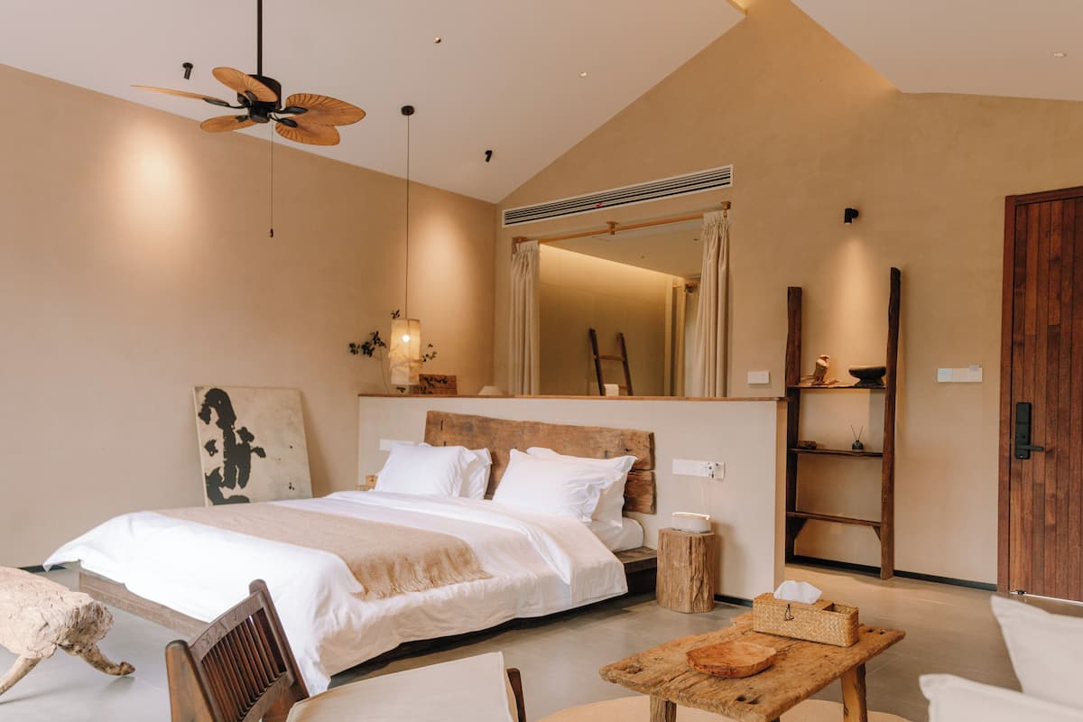 A modern bedroom with a large bed, a wooden headboard, a ceiling fan, a mirror, a wooden wardrobe, and a small wooden table with a bowl.