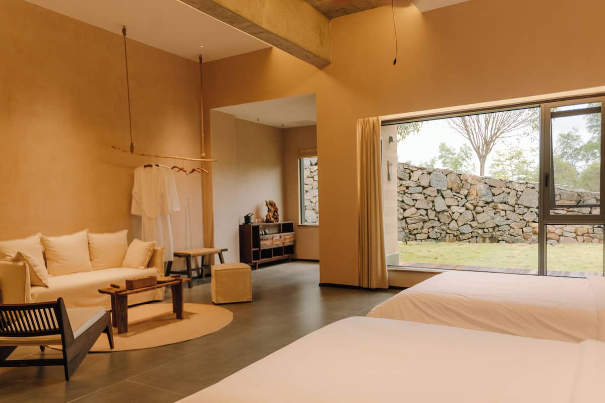 A spacious bedroom with a large bed, a sofa, and a small table. There is a view of a stone wall and greenery outside.