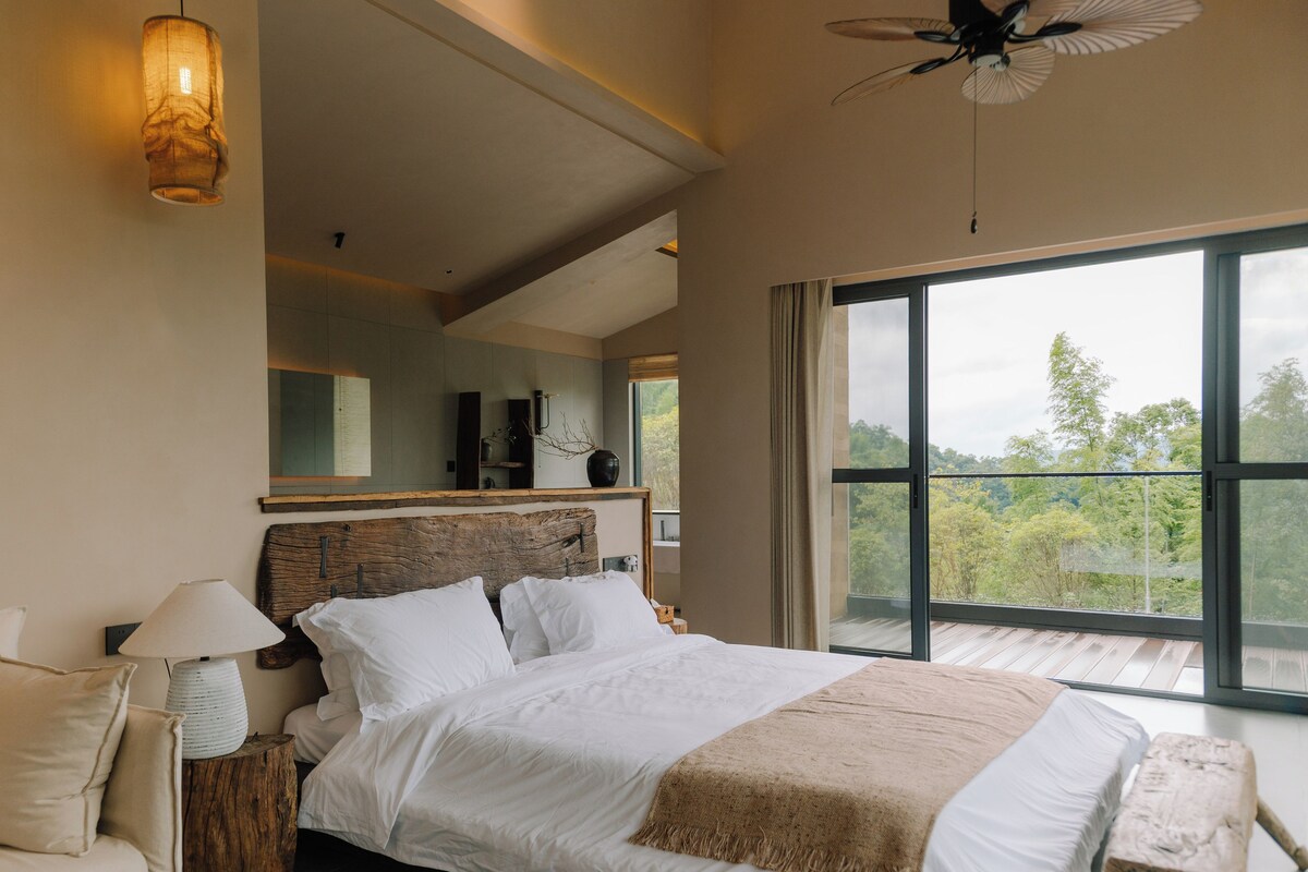 A bedroom with a large bed, a nightstand, a lamp, and a view of the outdoors through a large window.