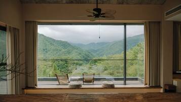 A room with a large window offering a view of lush green mountains. There are two wicker chairs and a small round table on the windowsill.