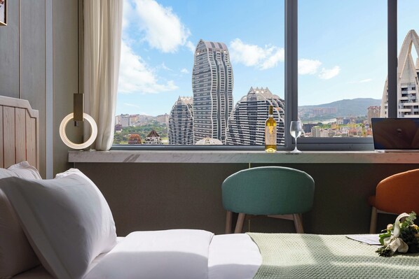 A hotel room with a bed, a desk with a laptop, a bottle, and a glass. A window offers a view of tall buildings and a cityscape.