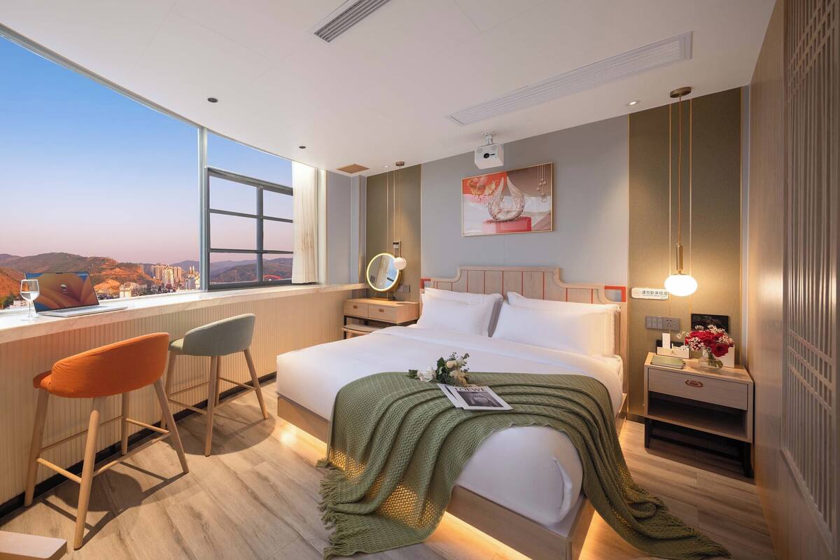 Moderni hotellihuone, jossa on suuri sänky, työpöytä kannettavalla tietokoneella, kaksi tuolia, yöpöytä lampun kanssa ja näkymä kaupungin ylle.