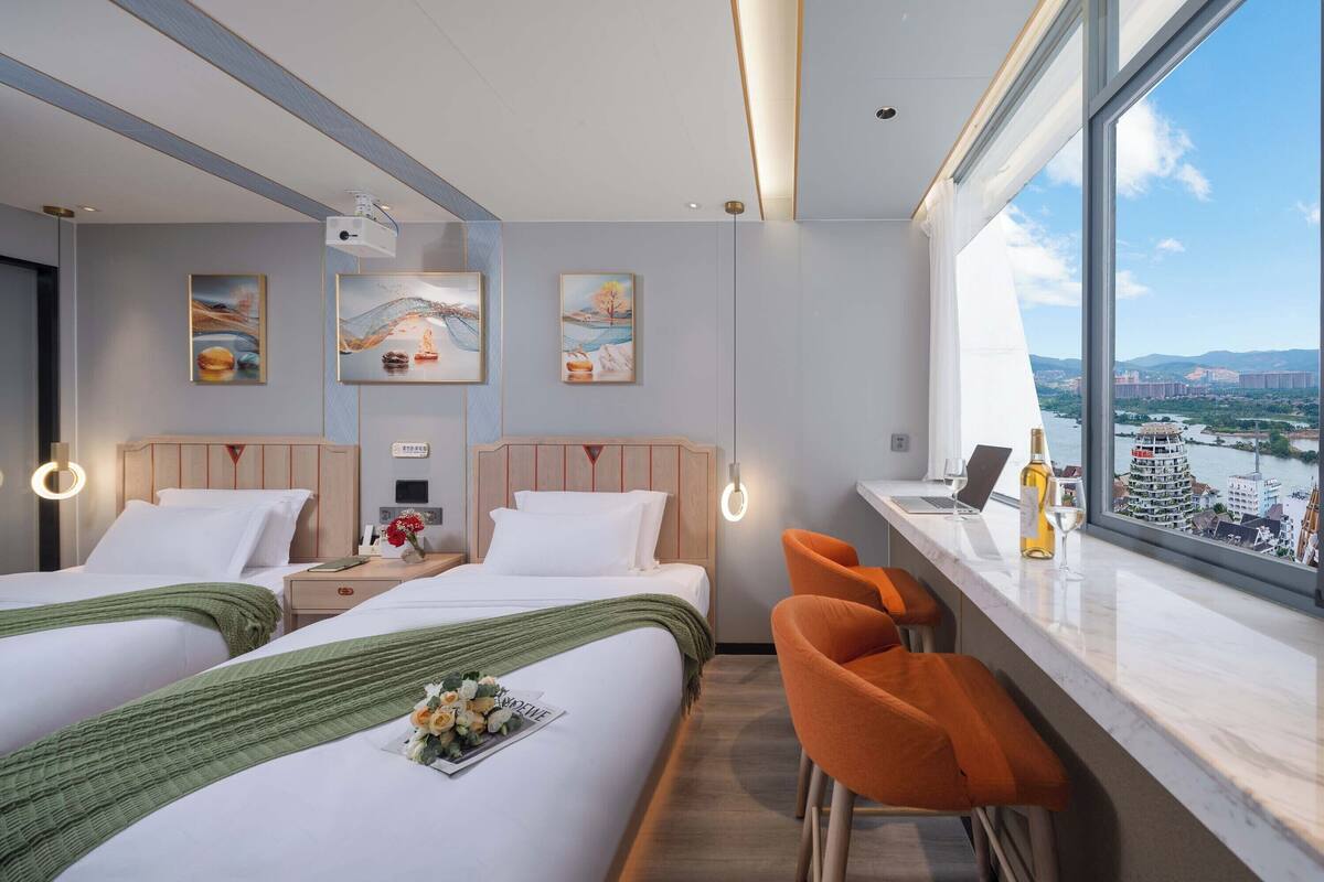 Hotellihuone, jossa on kaksi sänkyä, työpöytä kannettavalla tietokoneella, pullo viiniä ja näkymä kaupunkimaisemaan.
