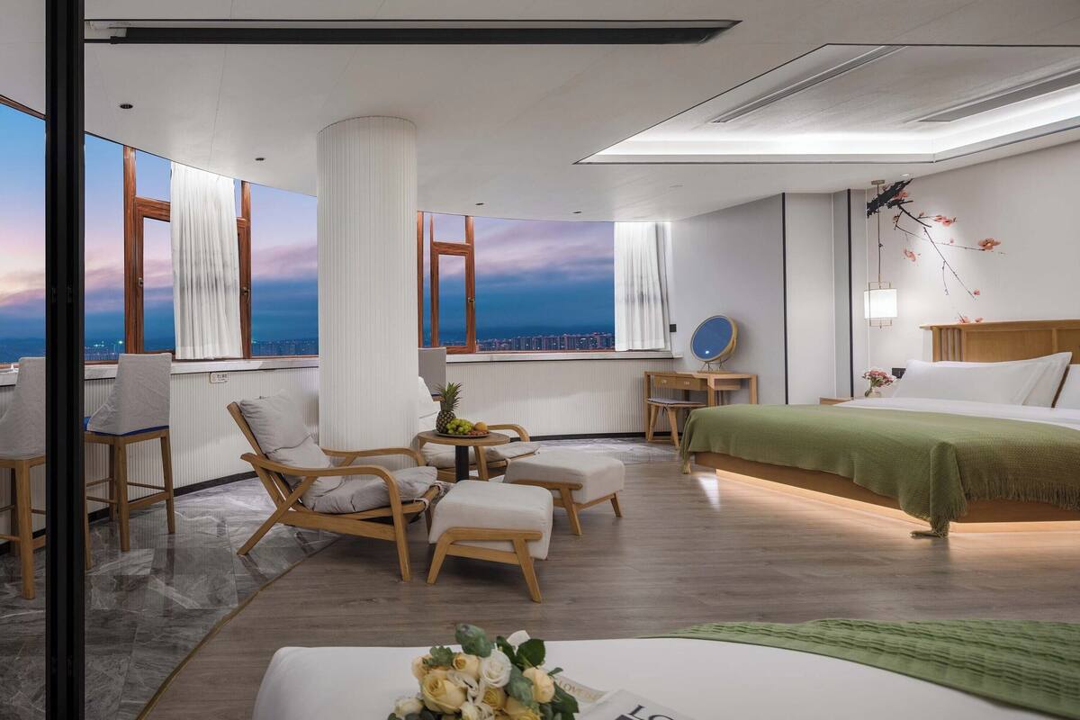 Moderni hotellihuone, jossa on suuri sänky, työpöytä ja oleskelualue tuoleineen. Suurista ikkunoista avautuu näkymä kaupunkiin.