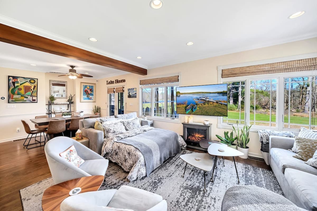 A spacious living room with a large TV, comfortable seating, a fireplace, and large windows with a lake view.