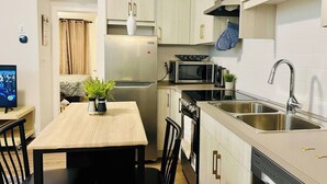 A compact kitchen with a refrigerator, microwave, stove, and sink. There is a dining table with chairs and a small potted plant.