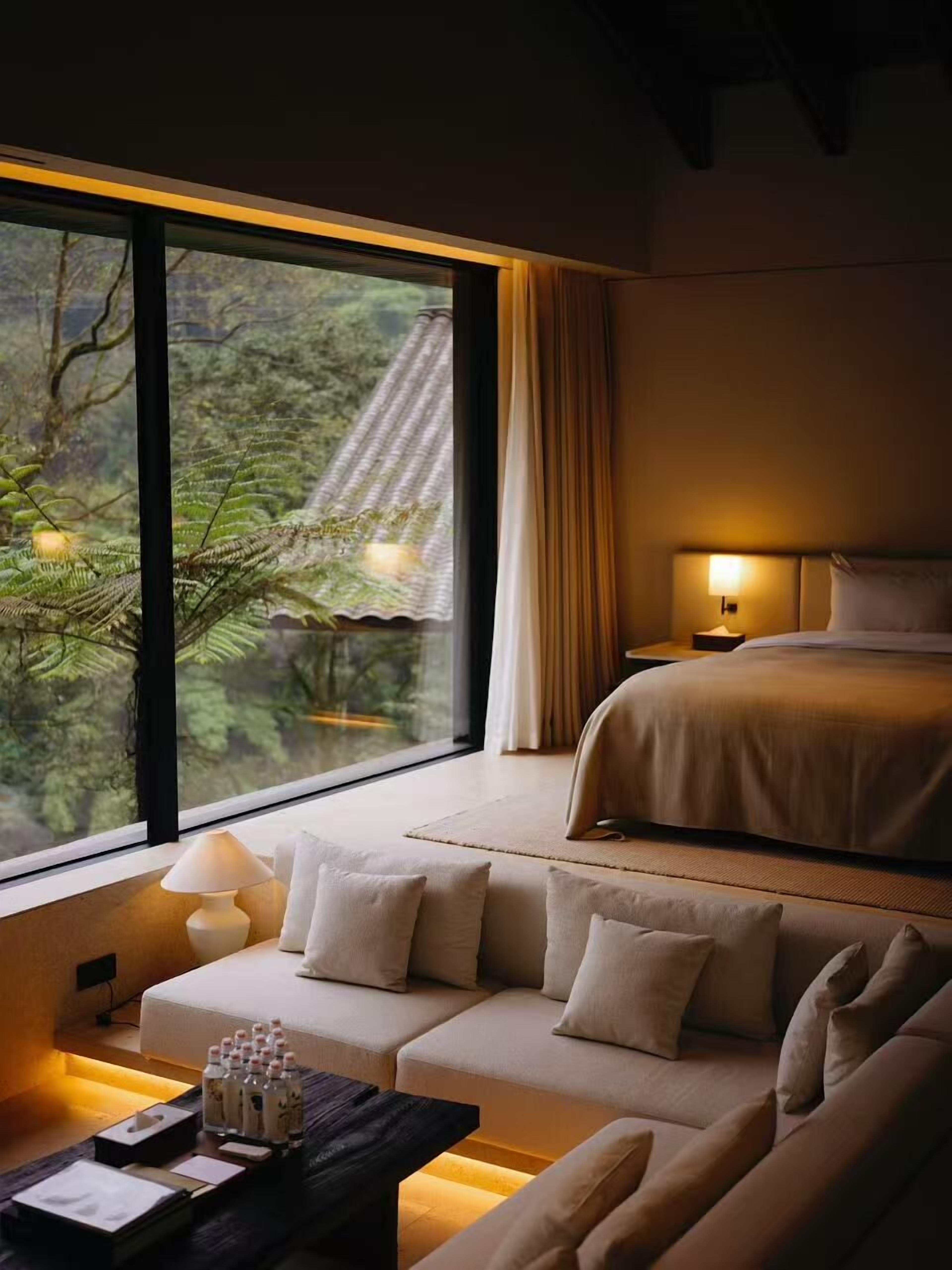 A modern bedroom with a large window, a sofa, a bed, and a bedside table with a lamp.