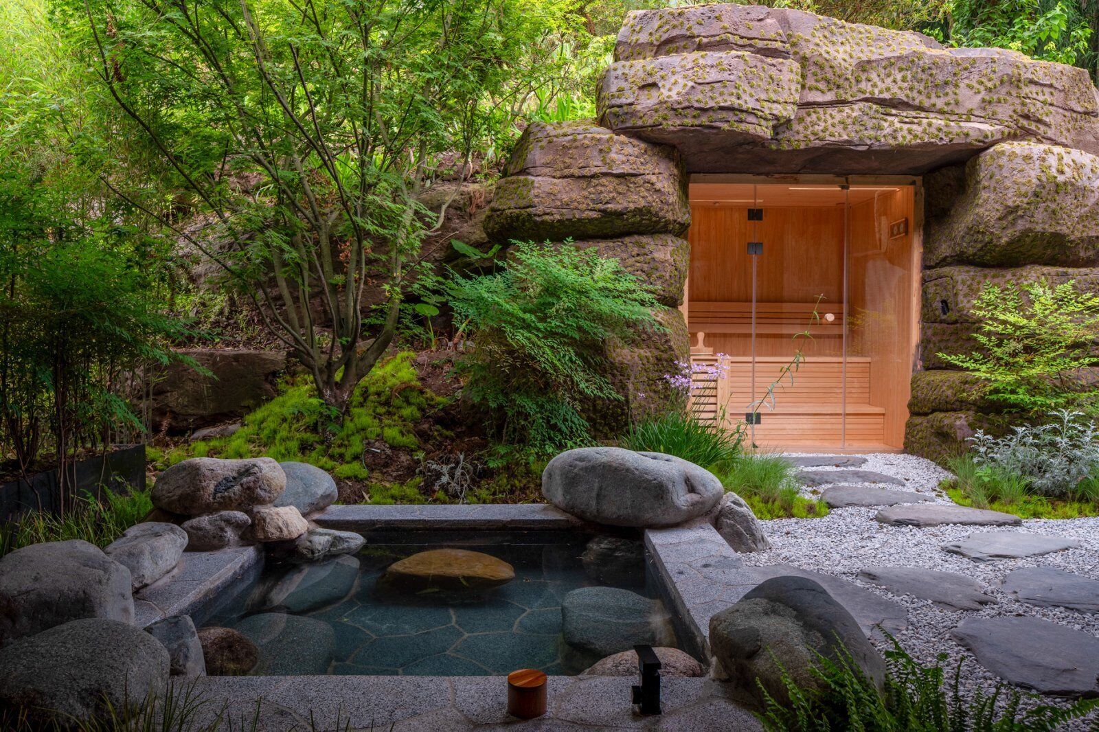 A traditional Japanese-style building with a wooden door, surrounded by lush greenery and a rock garden with a small pond.