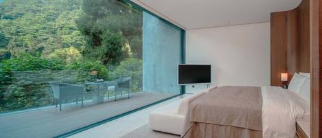 A modern bedroom with a large window, a bed, a TV, and a small seating area with a view of a forested landscape.