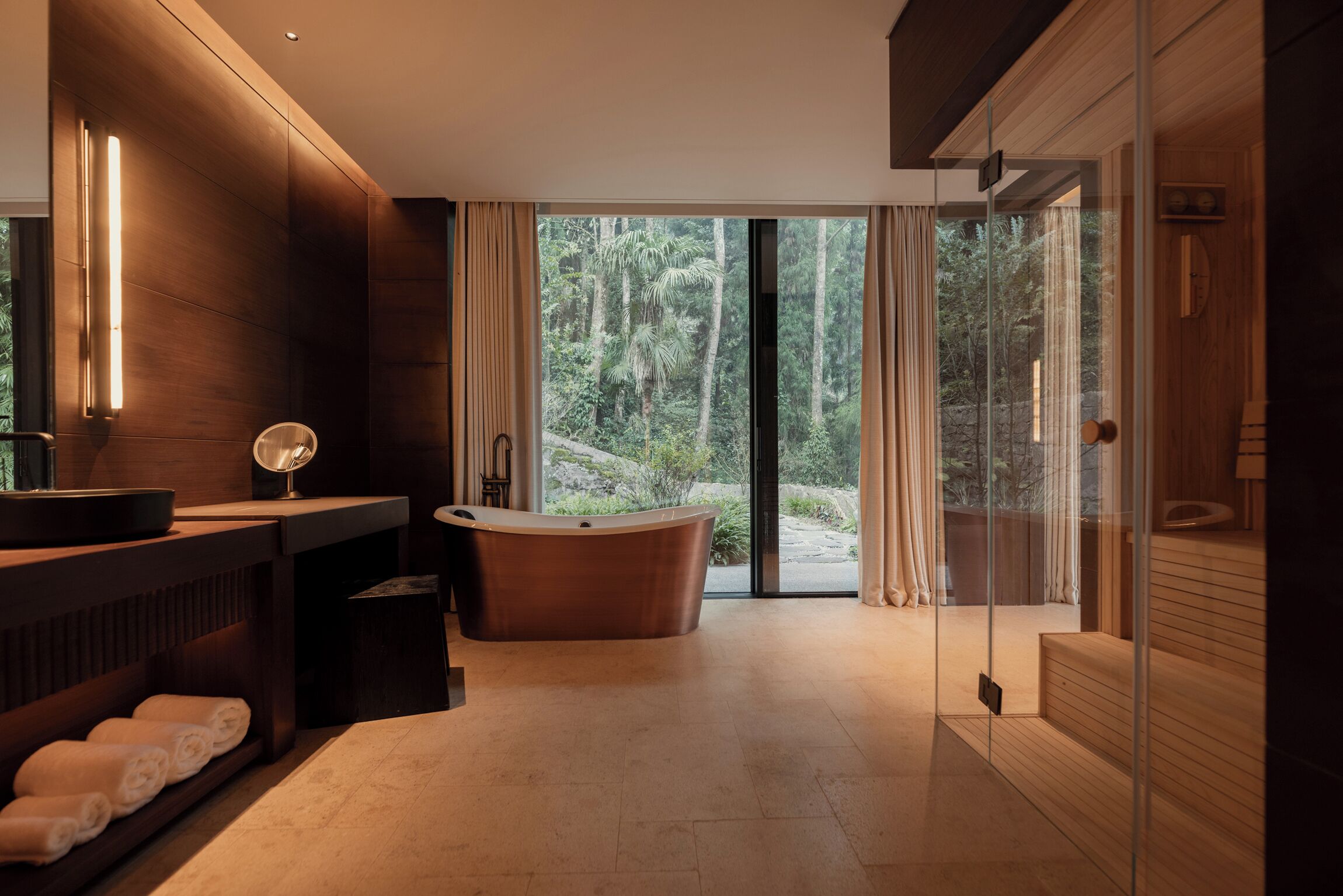 A modern bathroom with a freestanding bathtub, a vanity with a mirror, and a large window offering a view of a forested area.