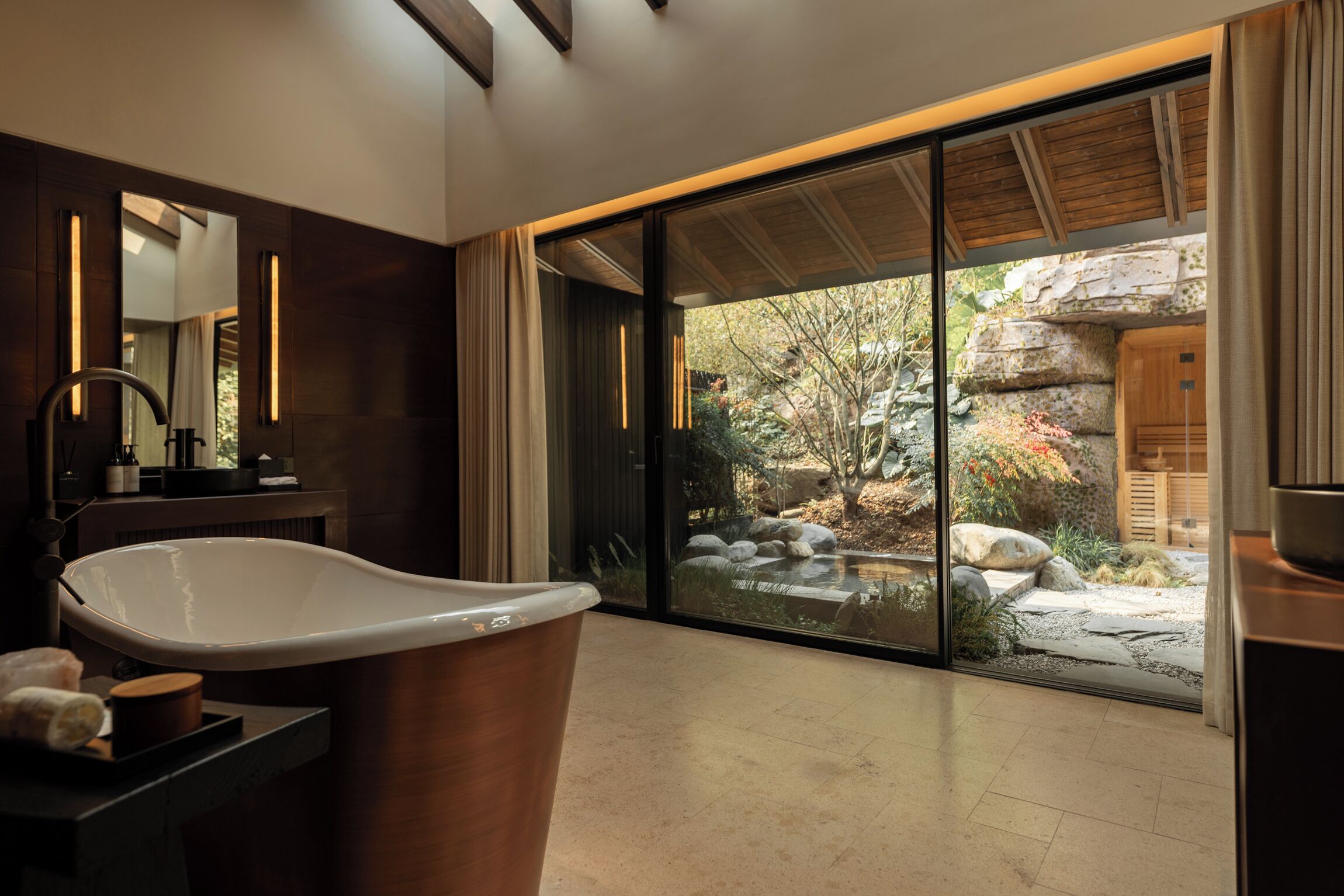 A modern bathroom with a freestanding bathtub, a large mirror, and a view of a landscaped garden through glass doors.