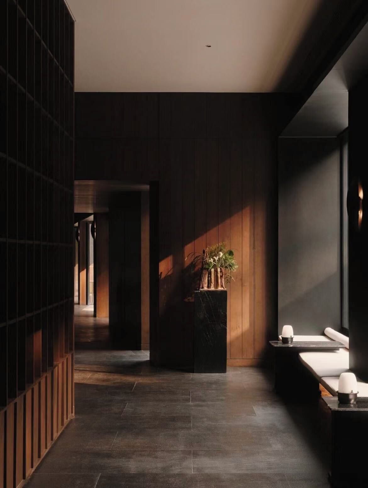 A modern interior with a dark wood panel wall, a central planter with greenery, and a seating area with a white chair and a small table.