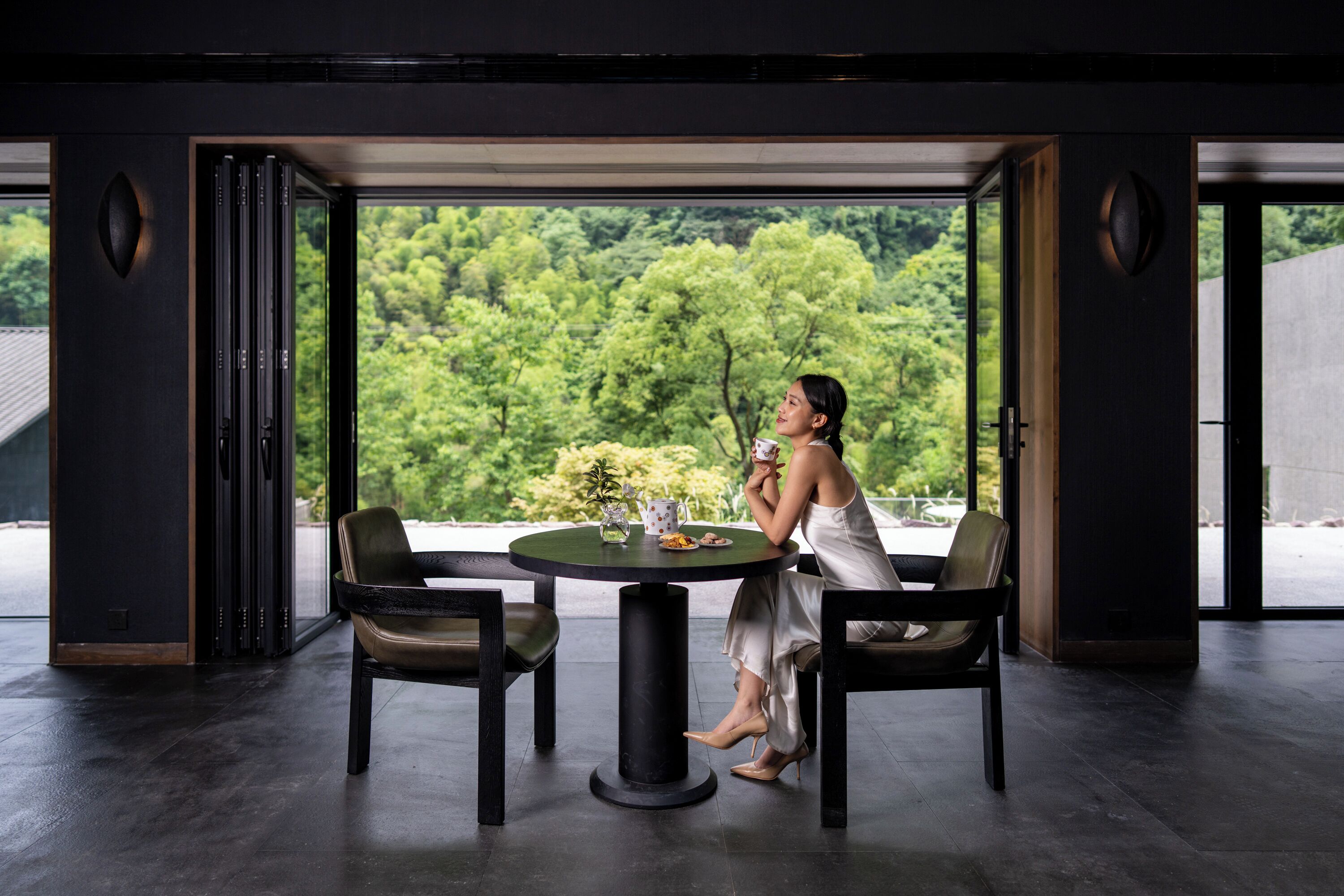 Un interior moderno con una mesa redonda, dos sillas y vistas a un paisaje verde exuberante a través de amplios ventanales.