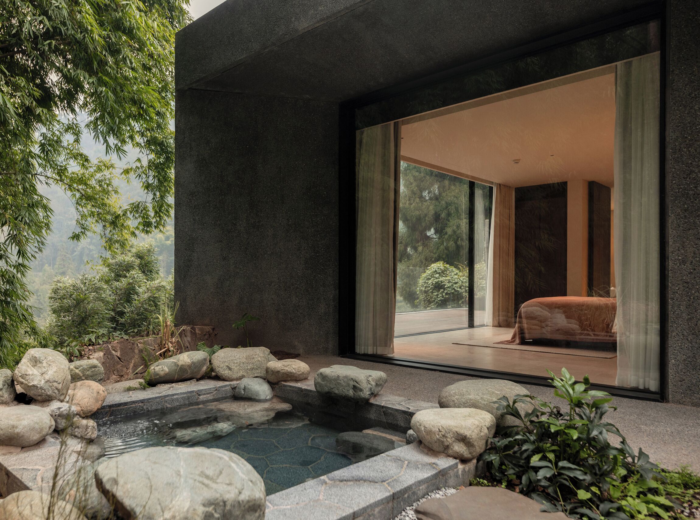 A modern outdoor space with a stone water feature, a seating area, and a view of a bedroom through large glass doors.