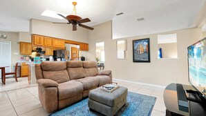 A living room with a brown sofa, a grey ottoman, a blue rug, a ceiling fan, and a flat-screen TV. - Spacious 3BR Home, Gym, Pool Table, Central Location (Deltona)