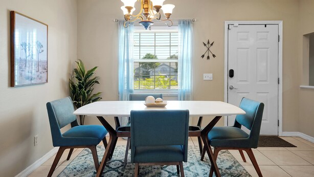A dining area with a white table, four blue chairs, a chandelier, a window with blinds, and a door. - Spacious 3BR Home, Gym, Pool Table, Central Location (Deltona)