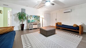 A living room with a ceiling fan, a large potted plant, a sofa, a coffee table, and a painting of a beach scene. - Cozy 2-bedroom apartment in centrally located Deltona. SLEEPS 7 PEOPLE! (Deltona)