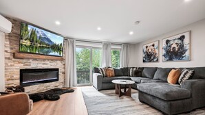 A modern living room with a fireplace, a grey sectional sofa, a coffee table, and a large flat-screen TV mounted on a brick wall.